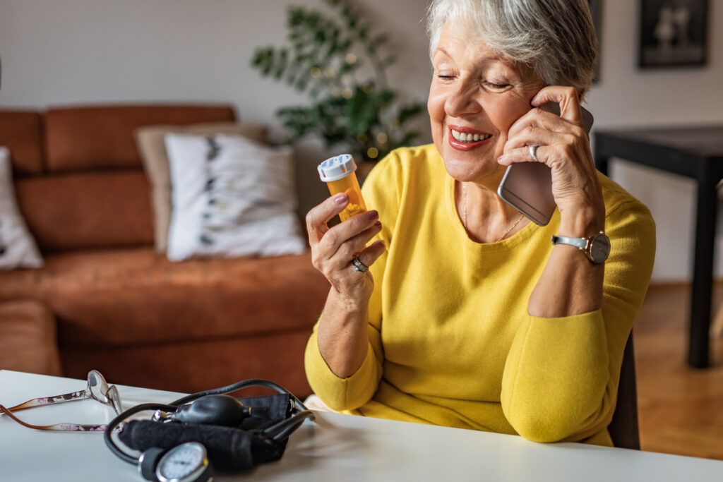 Photo of senior woman at home preparing to drink medical pill. She is holding a pill bottle and talking on the phone with a doctor
