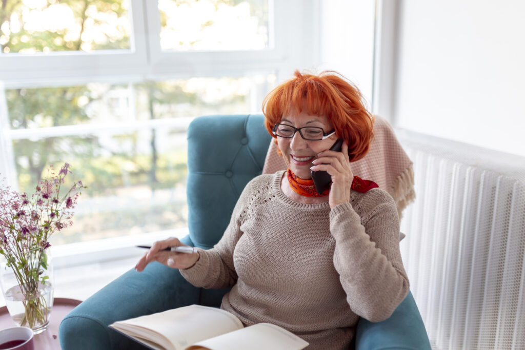 Senior woman with red hair sitting in an armchair, speaking on the phone and writing in a planner