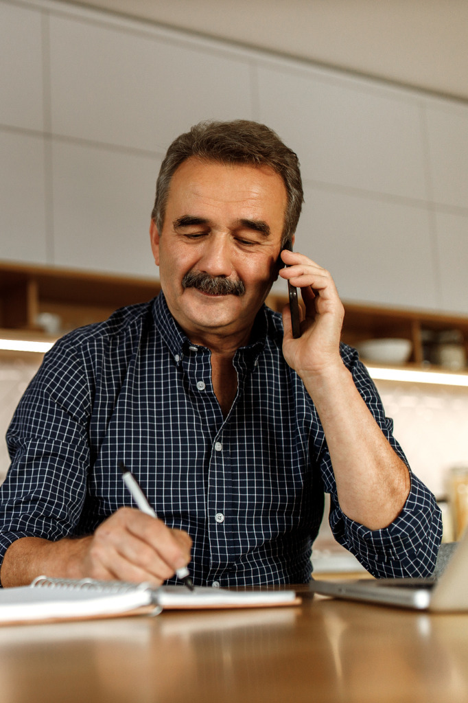 Mature man with mustache talking on phone and taking notes at kitchen table.