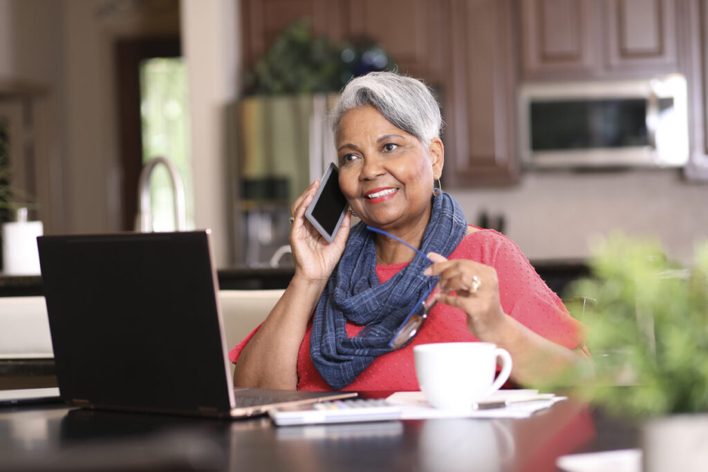 Senior woman in scarf at kitchen table talking on cell phone and using laptop.