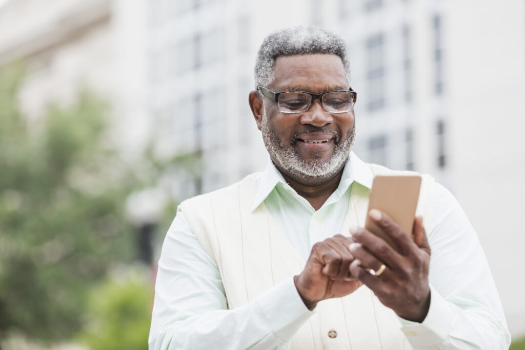 A senior man wearing a button down shirt, vest and eyeglasses, standing outdoors on a city street, looking down at his mobile phone.