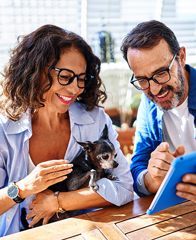 Mature Hispanic couple using touchpad sitting on table with dog at terrace