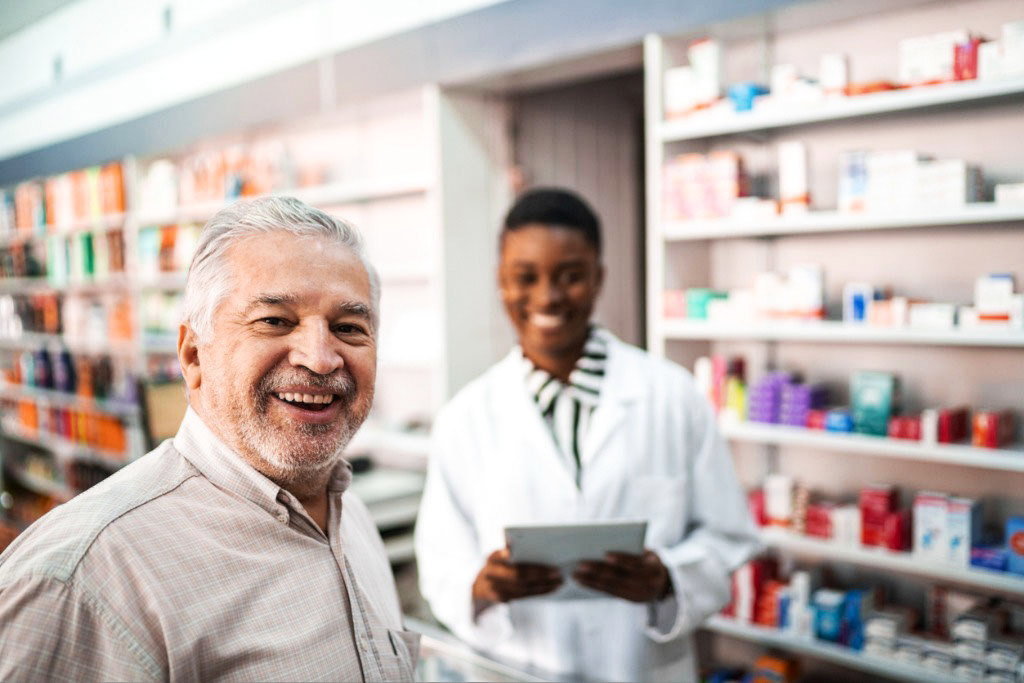 Senior man standing in a pharmacy and smiling next to his pharmacist.