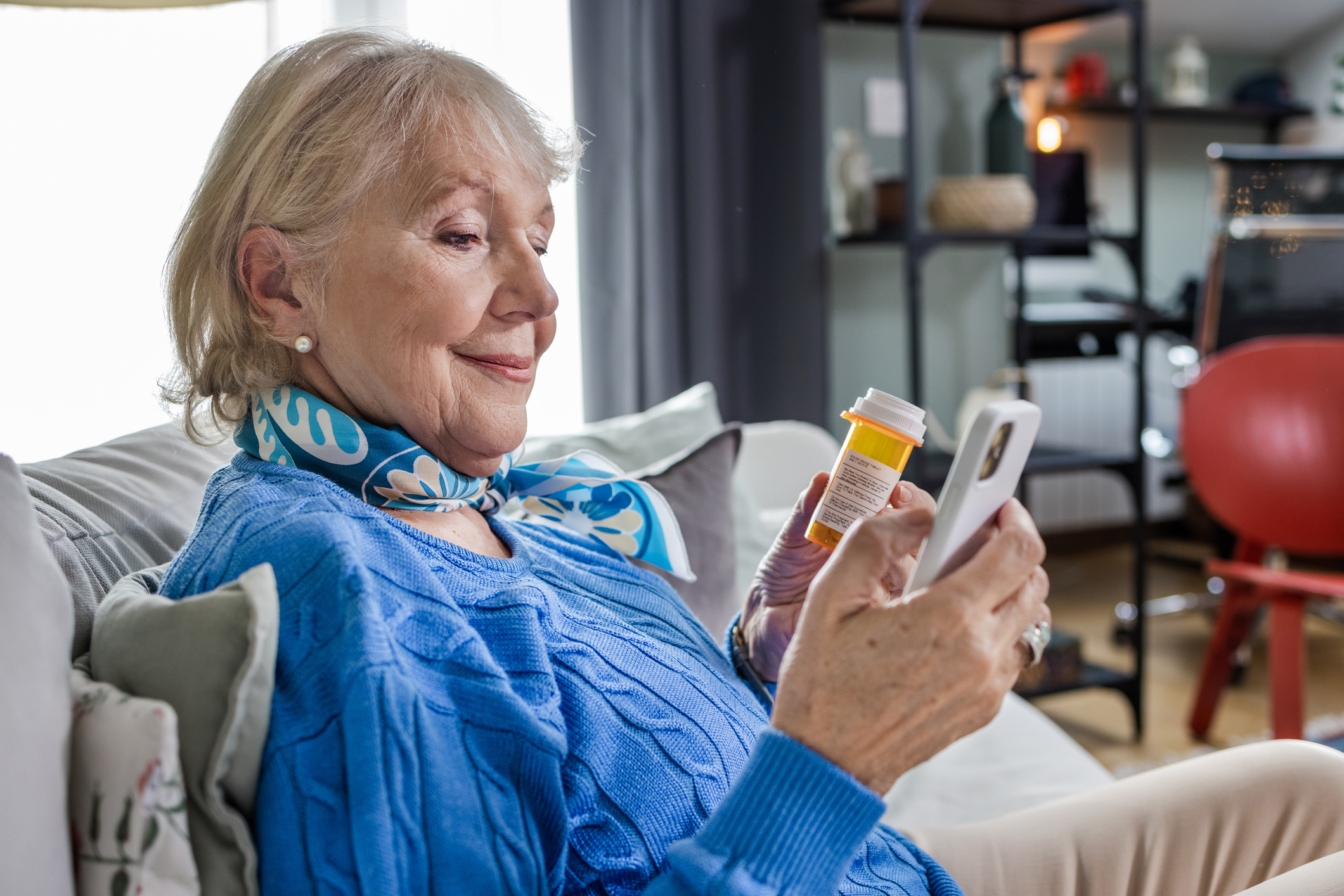 Photo a senior woman at home. She is holding a pill bottle and looking at a phone.
