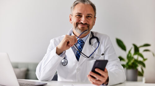 Portrait of a smiling doctor holding glasses and a mobile phone at the office.