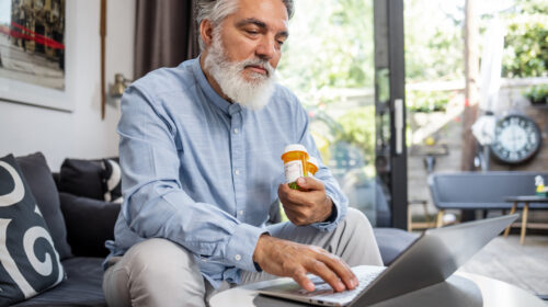 Photo of a senior man at home holding a pill bottle and using laptop. Health technology concept. stock photo
